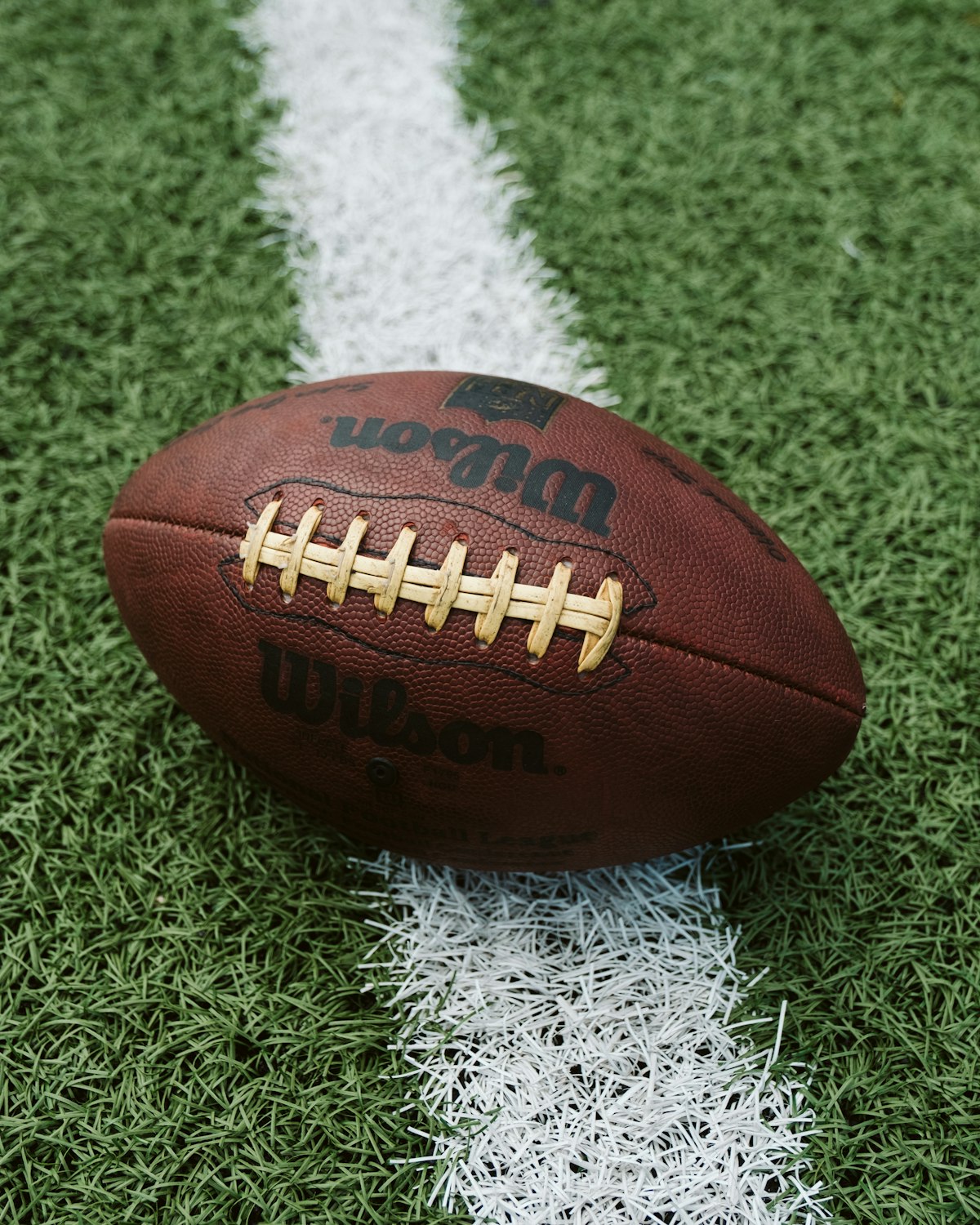 A brown leather American football with white laces resting on green grass next to a yard line