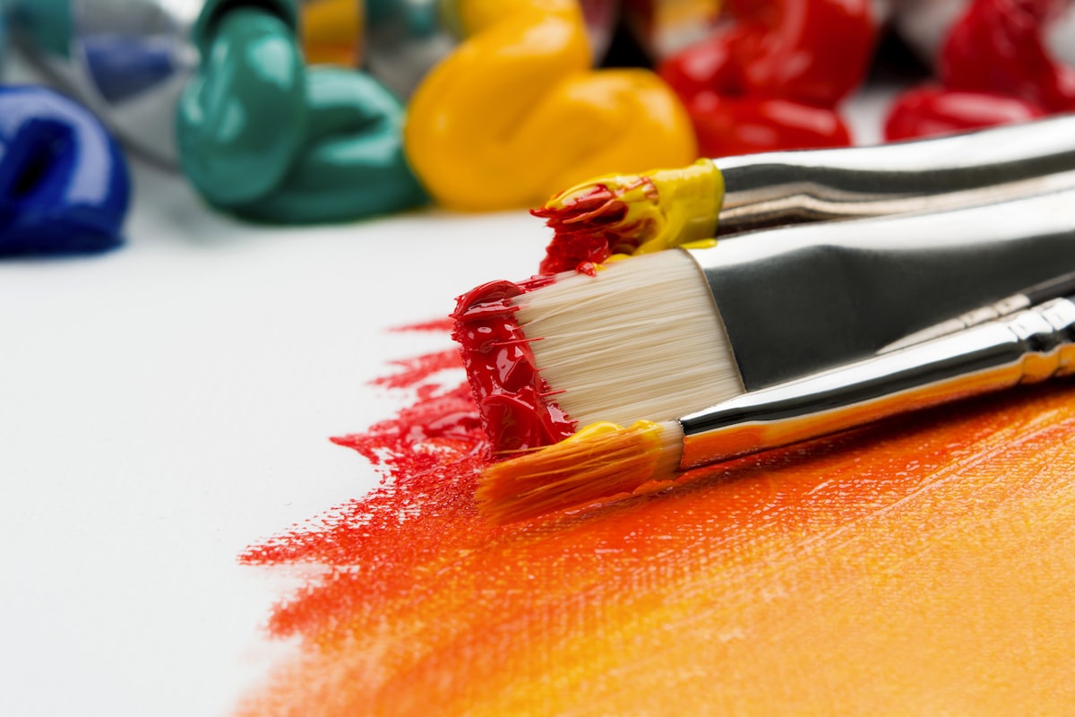 Two paintbrushes loaded with red and yellow paint resting on a half-painted canvas