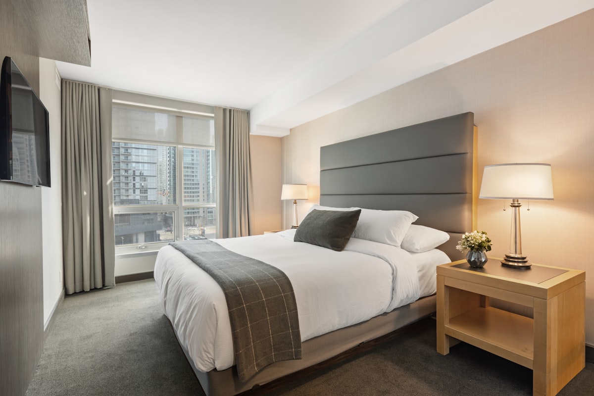 A neatly made bed in a sparse, sunlit room, white linen, simple and ordered